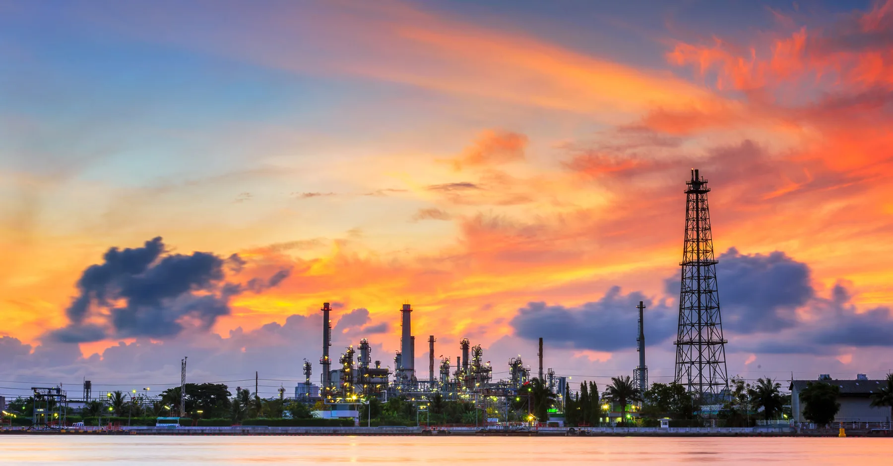 Oil refinery skyline at sunset with tall towers silhouetted against orange and pink sky beside a calm waterfront.